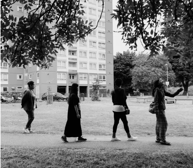 A group of people walking through a park, surrounded by trees and grass, with high-rise buildings visible in the background.