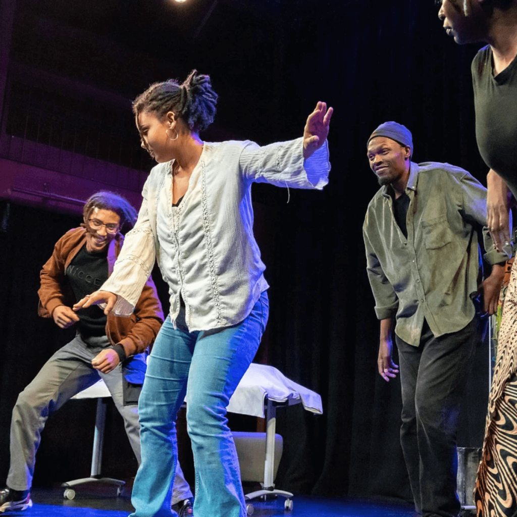 Mya Fraser, a mixed race women with tied back hair, a white blouse, and blue jeans, performs on stage as part of a play. Her head is turned away from the camera and she holds out her arms.