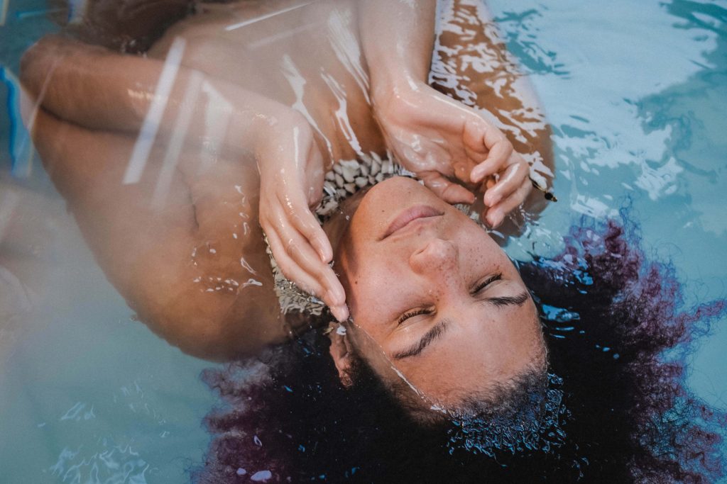 NGAIO, a mix raced woman with dark purple afro hair, lies in a pool of blue water. Her hands are cupped to her face and her eyes are closed.