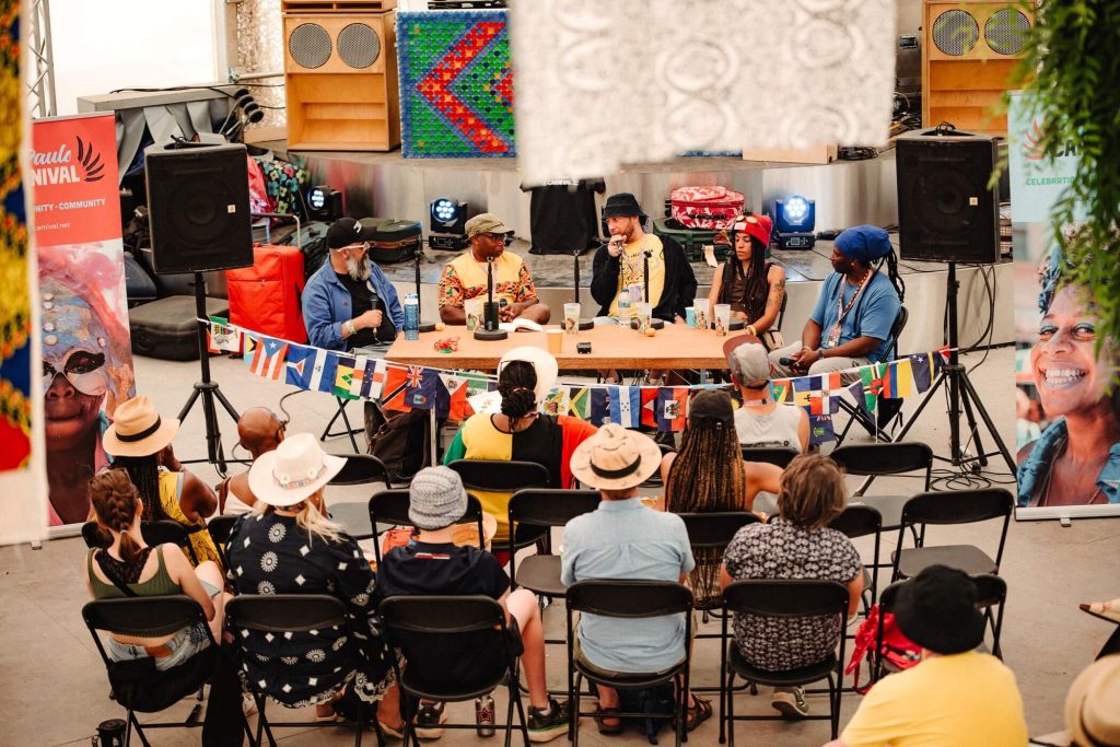 Five panel speakers sit at a desk, deep in discussion. In front of them and audience sit and listen. Around them are St Pauls Carnival banners and Caribbean flag bunting.