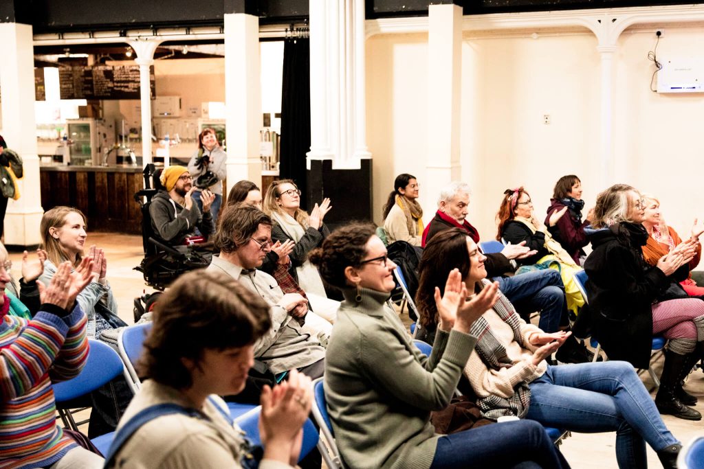 An audience sit in a hall, smiling and applauding what they are watching.