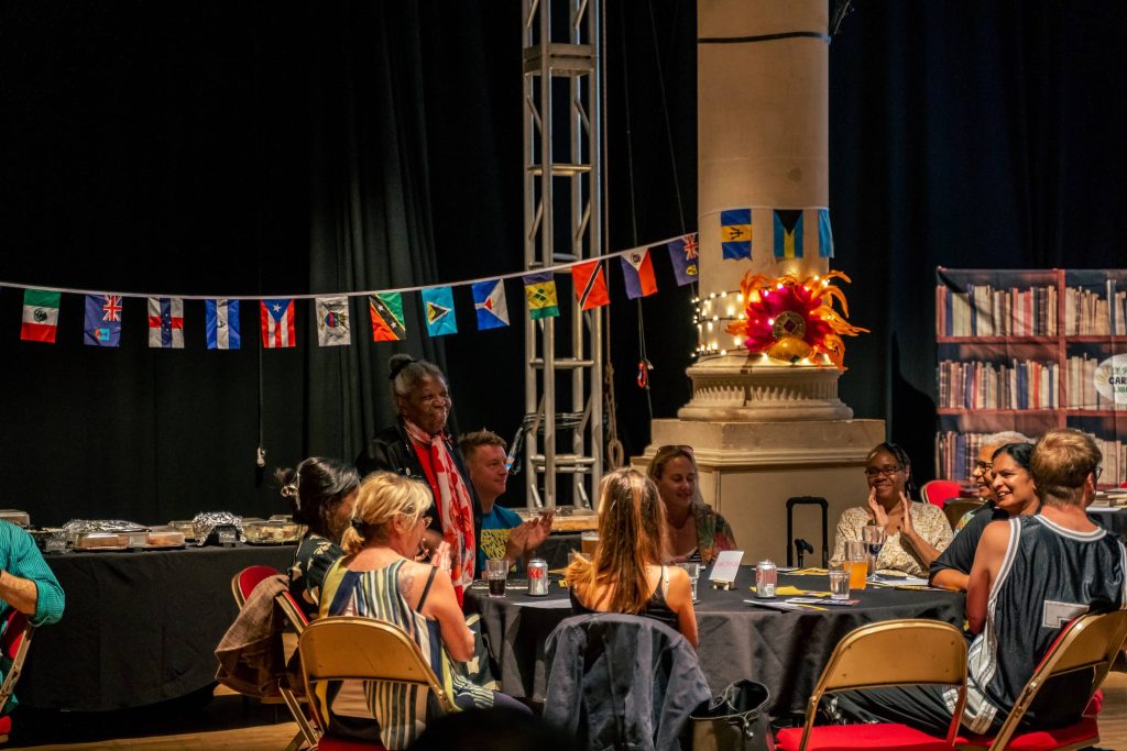 A group of people sit round a circular table, clapping. A black woman with grey hair is stood up, smiling. The space around them is decorated with Caribbean flags, fairy lights, and a Carnival headdress.