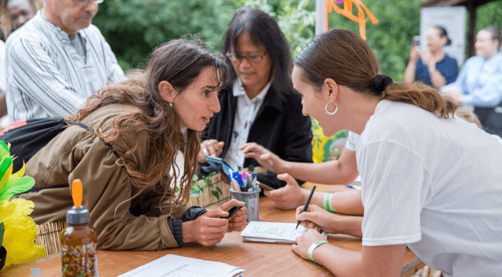 Two woman lean across a table towards one another. One woman has brown hair in a ponytail and wears a white t-shirt. She is writing on a notepad. The other women has long brown hair and wears a brown jacket.