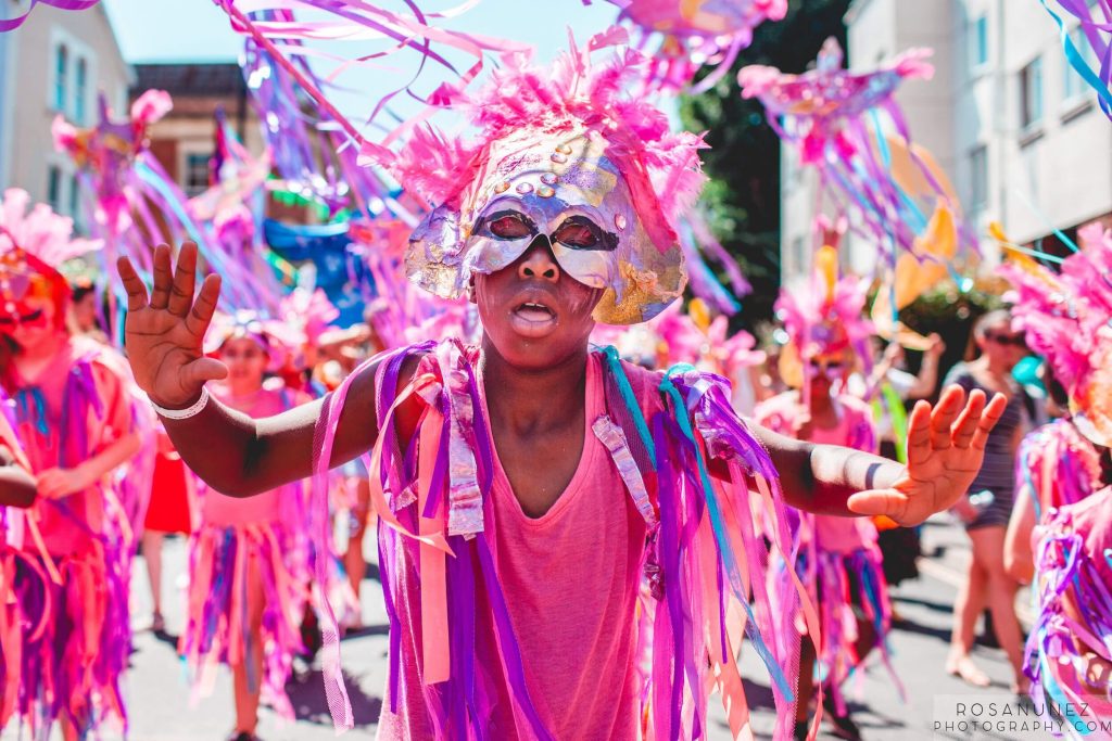 A woman dances as part of the St Pauls Carnival procession. She is wearing a pink dress with pink and purple ribbons on the shoulders, and a pink headdress. Her facial expression suggests she is totally lost in the moment. In the background, other people wearing the same costume also dance.