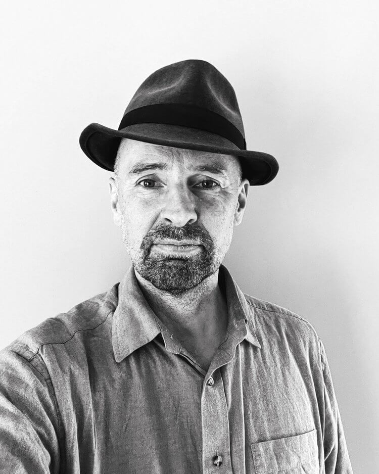 David Jubb, a middle aged man with a beard, looks directly down the camera. He wears a shirt and trilby hat.