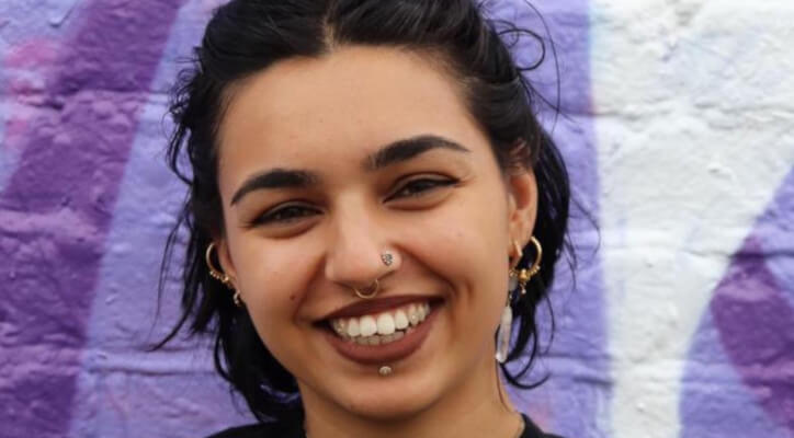 Anjali Prashar-Savoie smiles directly into the camera. She has short black hair, brown lipstick and facial piercings in her lip, nose and septum.