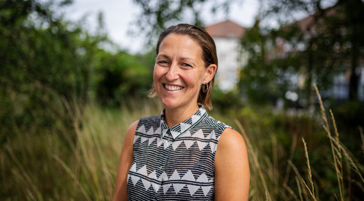 Martha Kind, a woman with short brown hair and wearing a black and white patterned blouse, smiles at the camera. She is outside. Behind her there is long grass and trees.