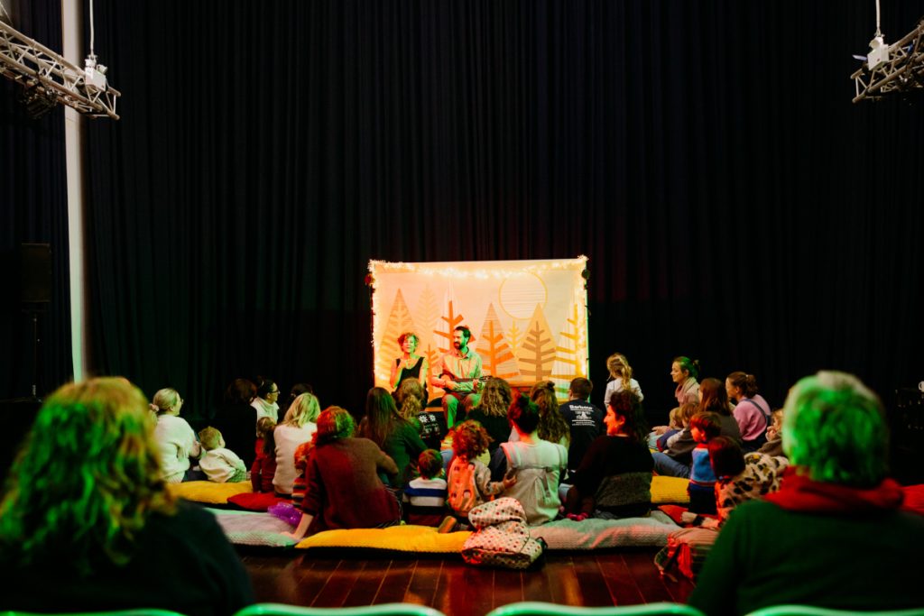 An image of a performance in a large hall. Audiences ranging from early years to parent/carers, sit on the floor and on cushions watching two performers in front of a projection screen, they are playing music instruments The lighting is soft and ambient