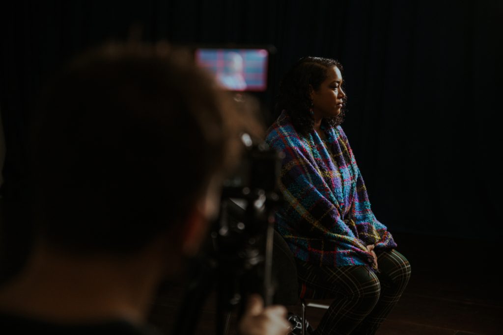 This is an image from the Art of Resistance project by Trinity Community arts and is a snap shot of a interview with Cleo Lake. Cleo lake sits on a chair in the back ground, a studio light with a diffuser box lights the front of her face. She is wearing a large shawl. In the foreground a camera opertative is filming Cleo