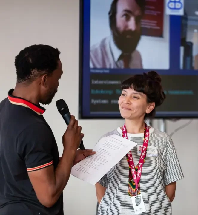 Maca Gomez-Gutierrrez is standing with her hands behind her back. She wears a black and white striped t-shirt, and has brown hair in a bun with a short fringe. She is smiling at a man who is facing her. In one hand, he holds a microphone that he is speaking into; in the other hand, he holds a piece of paper.