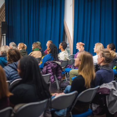 An image of audiences sitting on plastic chairs in a large hall. The image of the back of peoples heads and they are watching an unseen event infront of them. There is a range of people, ages, hair styles and backgrounds